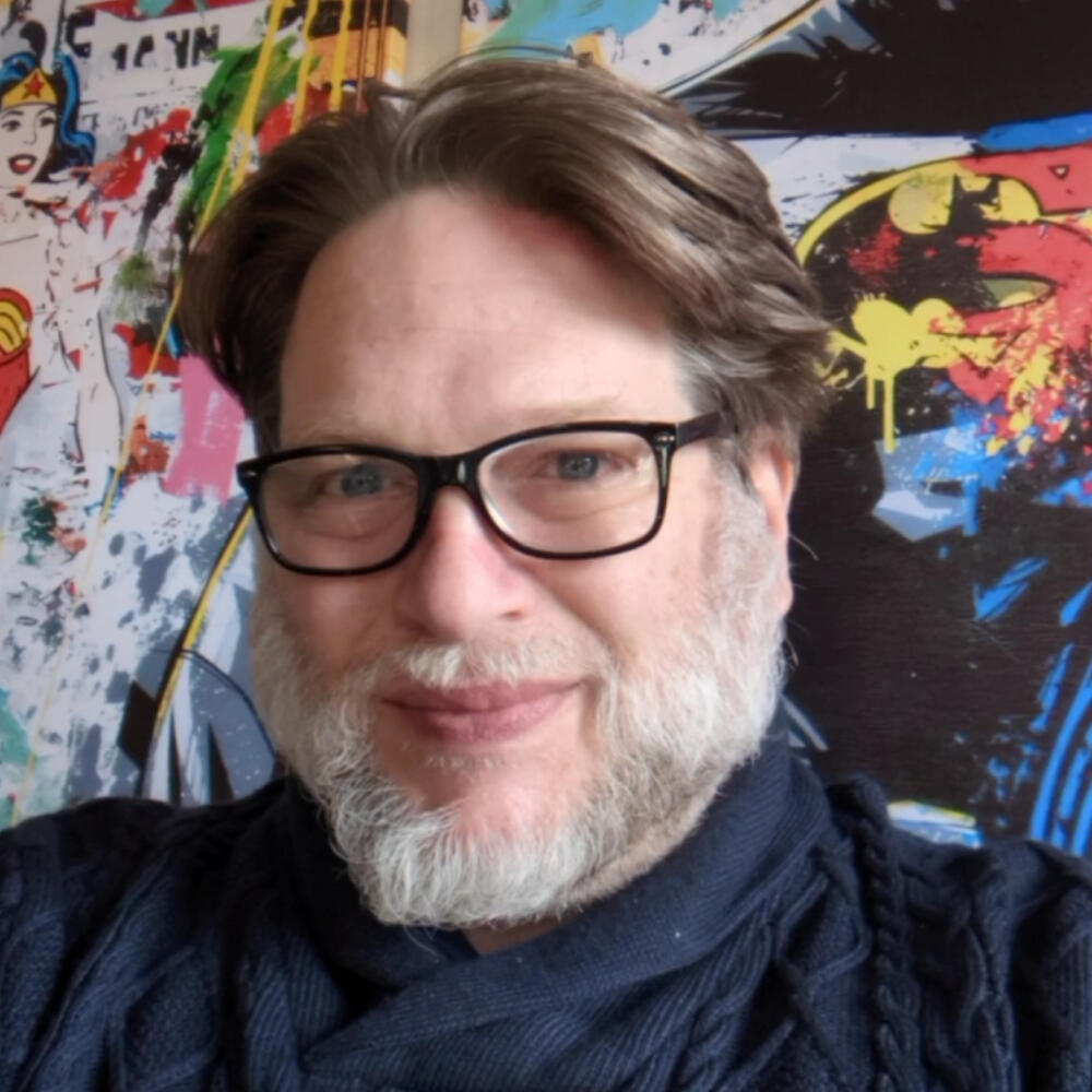 Chris Brogan with short hair, wearing glasses and a blue sweater. His background shows marvel artwork.