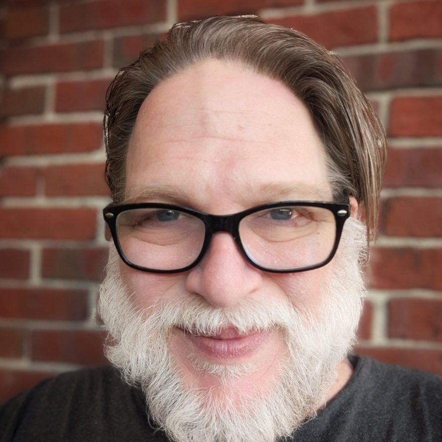 Portrait of Chris Brogan, smiling, with glasses and a white beard, in front of a brick wall